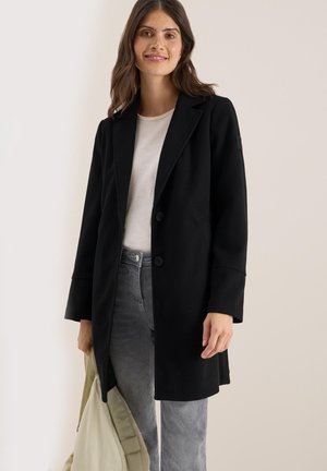 Manteau noir au design ajusté, deux boutons, revers crantés et poches latérales. Porté par-dessus un haut clair avec un jean gris et un sac.
