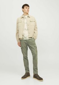 Chaqueta de mezclilla beis sobre una camiseta de tie-dye verde claro, combinada con pantalones cargo olivo y botas de senderismo marrones. Fondo liso, pose de pie.