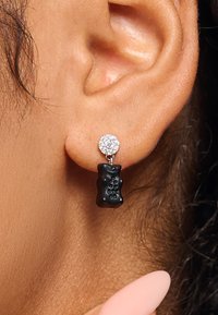 Schwarzer Ohrring in Form eines Gummibärchens mit einem funkelnden, runden Kristallstecker darüber. Das Metallbeschlag ist silberfarben und weist glatte Oberflächen auf.