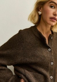 Brązowy dziany kardigan z okrągłym dekoltem, z dużymi szarymi guzikami na froncie i miękką teksturą, noszony na jasnej bluzce.