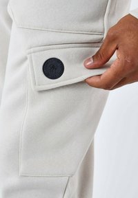 Los pantalones de chándal grises claros presentan un bolsillo lateral con un parche de logo redondo negro. La tela es suave y muestra detalles de costura visibles.