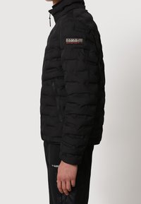 Chaqueta acolchada negra con cuello alto, bolsillo lateral con cremallera, textura acolchada y parche de logo en el brazo izquierdo.