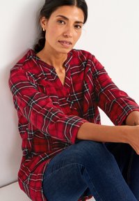 Rød ternet skjorte med knapper, sorte og hvide accenter, langærmet og med V-hals, båret med mørkeblå jeans, mod en hvid baggrund.