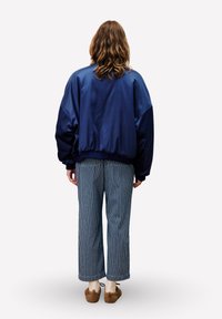 Marineblauwe oversized bomberjack met satijnen textuur, gecombineerd met wijde gestreepte broeken. Model staat met de rug naar de camera en draagt bruine schoenen.