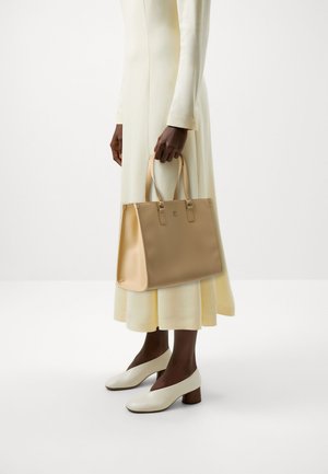 Borsa tote beige con manici doppi, texture liscia e hardware argento, tenuta da una persona che indossa un lungo vestito giallo pallido e scarpe bianche.