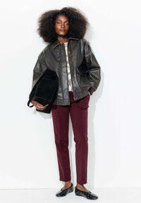 Veste en cuir noir au design structuré, pantalon bordeaux, et chaussures noires à enfiler avec des clous blancs, accompagnées d'un sac noir.