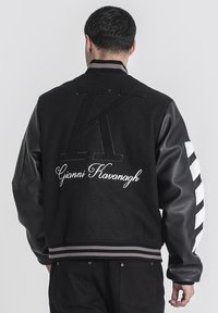 Čierna varsity bunda s vlnenným telom, koženými rukávmi, embosovaným „K“ a „Gianni Kavanagh“ na chrbte, rebrovaným golierom a lemom. Pruhované akcenty.