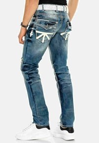 Jean en denim bleu à coupe slim, avec des zones usées, des designs uniques de poche arrière et une ceinture à motifs blancs.