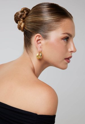 Goldener Creolen-Ohrring mit geschwungenem Design und polierter Metalloberfläche. Das Haar ist zu einem ordentlichen Dutt gestylt, sodass der Ohrring sichtbar ist.