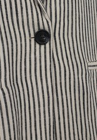 Bomullsväst med svartvita vertikala ränder, med en enda svart knapp och en ficka på vänster sida. Strukturerad tyg.