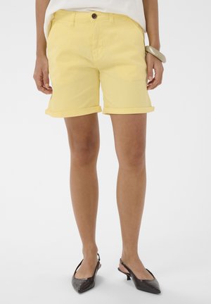 Žena v žltých kraťasoch s vyhrnutým lemom, bielom topu, hnedých nízkych špicatých sandáloch na zadnej časti nohy a veľkom béžovom náramku na ľavom zápästí.