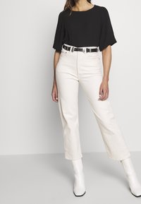 Černý top s krátkým rukávem spojený s krémovými kalhotami s vysokým pasem a úzkým, zdobeným černým opaskem. Bílé kotníkové boty na podpatku doplňují celkový vzhled.