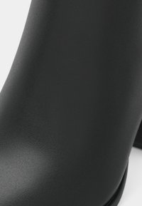 Μαύρη μπότες στον αστράγαλο με λεία, ματ φινίρισμα. Ελαφρώς κωνική σιλουέτα και τετράγωνη ψηλή σόλα. Χωρίς ορατά μεταλλικά στοιχεία ή σχέδια.