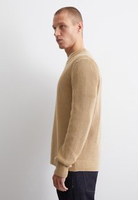 Beige ribbed Strickpullover mit langen Ärmeln, Rundhalsausschnitt und strukturierten Details, kombiniert mit dunklen Jeans. Seitenansicht.