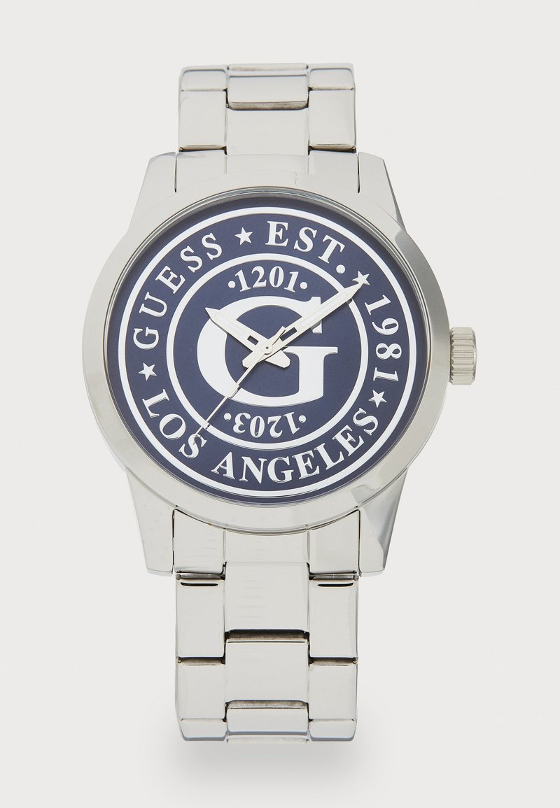 Reloj de metal plateado con una esfera redonda en color navy. Presenta un gran logotipo G en blanco, manecillas plateadas y una correa de metal con eslabones pulidos.