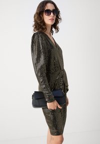 Robe à sequins noire et or avec des manches longues et un décolleté en V profond ; accompagnée d'un sac à main noir à rabat. Texture lisse.