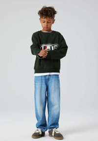 Zielona bluza z grafiką retro telewizora; jasnoniebieskie, luźne dżinsy; brązowe buty. Tkanina wydaje się miękka, z luźnym krojem.