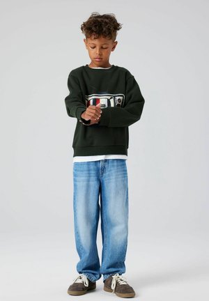 Zelená mikina s retro grafikou televizoru; světle modré oversized džíny; hnědé boty. Tkanina působí měkce a volně.