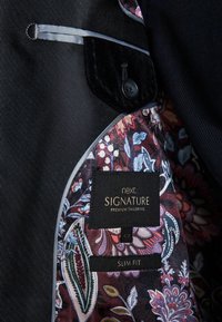 Interior de chaqueta de traje negra con forro de estampado floral en burdeos, azul y crema, con una etiqueta "next SIGNATURE" y un detalle de botón.