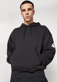 Junger Mann mit kurzen Haaren trägt einen schwarzen Hoodie mit einer Fronttasche und einem weißen Logo auf dem linken Ärmel, steht vor einem schlichten Hintergrund.