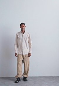 Bărbat stă în fața unui perete gri simplu, purtând o cămașă deschis bej cu nasturi, pantaloni cargo largi de culoare khaki și pantofi negri de birou, privind spre înainte.