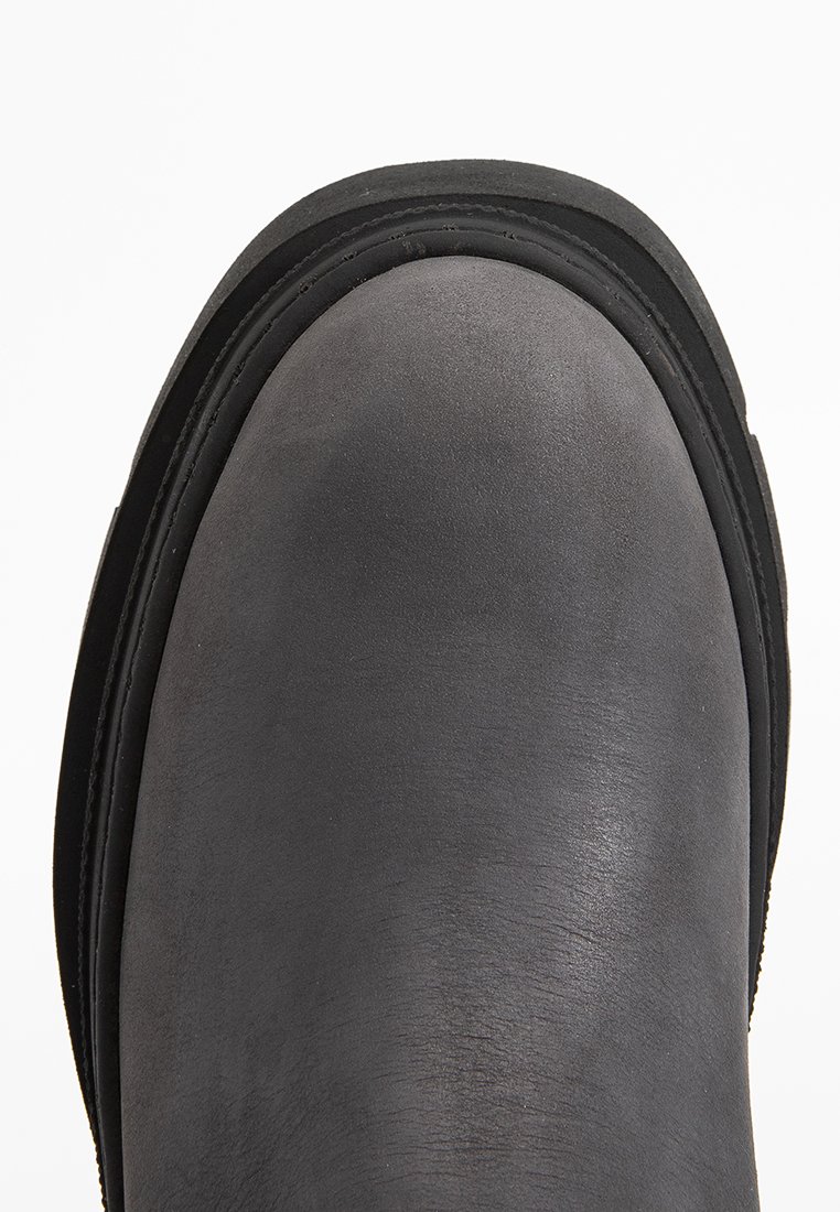 Nahaufnahme der abgerundeten Spitze eines schwarzen Lederstiefels mit sichtbaren Nähten und einer dicken Gummisohle vor einem weißen Hintergrund.