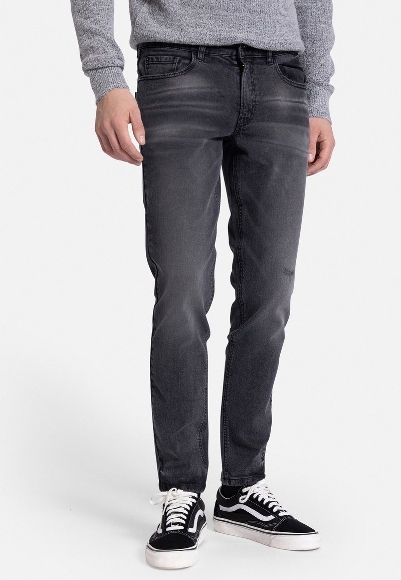 Homme portant un jean noir slim délavé, un pull gris et des baskets noires et blanches, debout devant un fond blanc.