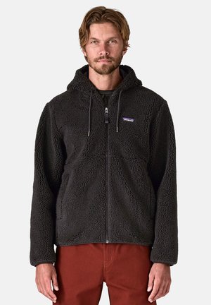 Felpa con cappuccio in pile nera, dotata di zip frontale, cappuccio regolabile e materiale testurizzato. Il modello indossa pantaloni arancione-marroni.