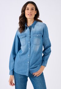 Camicia in denim azzurro con bottone frontale, due tasche sul petto e maniche lunghe. Caratterizzata da colletto classico e orlo leggermente curvato.