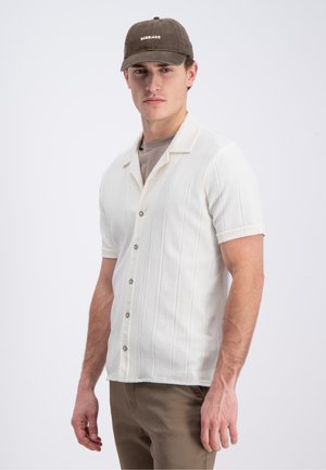 Mann trägt ein weißes kurzärmeliges Hemd mit strukturierten vertikalen Streifen, braune Hose und eine dunkelbraune Gabbiano-Mütze.