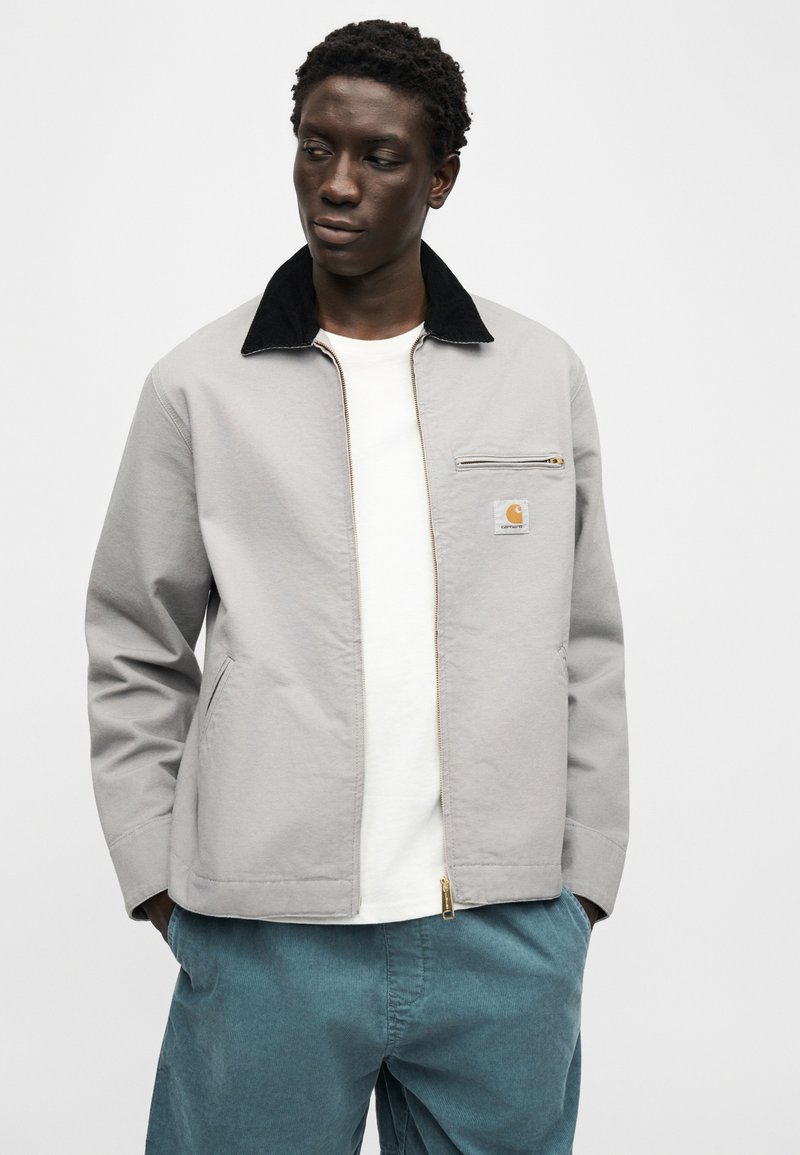 Homme portant une veste zippée gris clair avec col noir, chemise blanche et pantalon turquoise, debout les mains dans les poches, regardant à gauche.