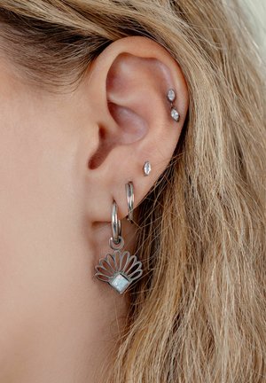 Nahaufnahme eines blonden Ohrs mit vier silbernen Ohrringen, darunter ein Creolen mit einem fächerförmigen Anhänger, der einen quadratischen Opalstein zeigt.