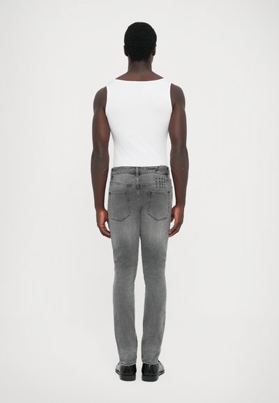Graue Jeans im Slim-Fit-Stil mit Gesäßtaschen und dezenten Steppnähten, kombiniert mit einem weißen ärmellosen Oberteil und schwarzen Schuhen.
