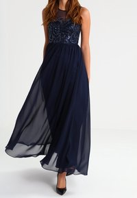 Mörkblå maxiklänning med en figurnära bärnstensdekorerad överdel och genomskinlig överdel, med en flödande kjol och utan ärmar. Svarta klackar kompletterar looken.