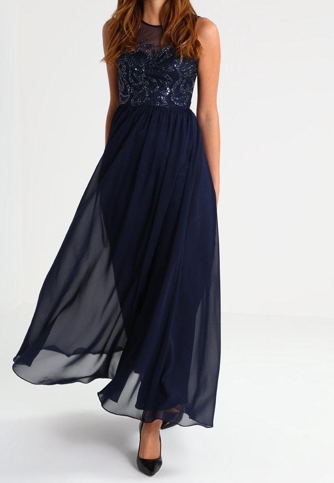 Mörkblå maxiklänning med en figurnära bärnstensdekorerad överdel och genomskinlig överdel, med en flödande kjol och utan ärmar. Svarta klackar kompletterar looken.