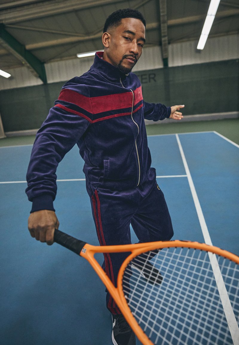 Marineblå velour treningsdrakt med røde striper, glidelåsjakke og smale bukser. Modellen holder en oransje tennisracket på en blå bane.
