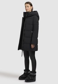 Fekete puffkabát kapucnival, steppelt mintázattal, oldalsó cipzáras zsebbel, fekete leggingsszel és masszív fekete csizmákkal párosítva.