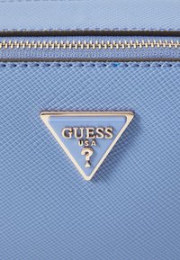 Materiale testurizzato blu chiaro con una zip argentata. Presenta un logo triangolare blu e argento con la scritta "GUESS USA" e un simbolo di accento.