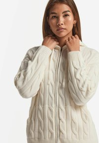 Vit kabelstickad huvtröja med dragkedja, ribbade ärmmuddar och sidofickor. Mjuk textur och avslappnad passform, lämplig för lagerpåklädnad.