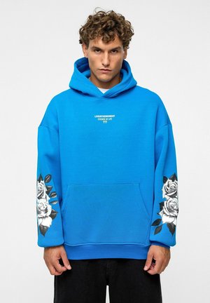 Blauwe hoodie met een voorzak, met witte tekst op de borst en zwarte rozenafbeeldingen op de mouwen. De tekst luidt "LIVEINTHEMOMENT ESSENCE OF LIFE 777."