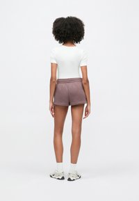 Γυναίκα με σγουρά μαλλιά που στέκεται γυρισμένη προς τα πίσω, φοράει λευκή μπλούζα με κοντά μανίκια, μωβ σορτς, λευκές κάλτσες και λευκά αθλητικά παπούτσια σε απλό φόντο.