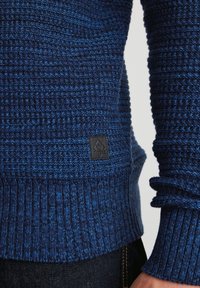 Dunkelblauer gestrickter Pullover mit einem strukturierten Muster, gerippten Bündchen und einem kleinen schwarzen Logopatch near dem Saum. Weiches, mittleres Material.