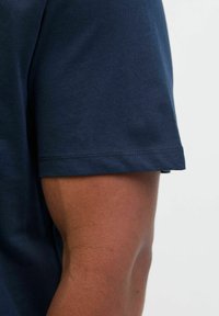Nahaufnahme eines dunkelhäutigen Arms, der ein dunkelblaues kurzärmliges Hemd trägt, vor einem einfachen hellen Hintergrund.
