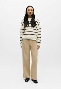 Megztinis su kremine baze ir tamsiai rudomis juostelėmis, derinamas su smėlio spalvos plačiomis kelnėmis. Medžiaga yra minkšta; megztinis turi raištelį apačioje.