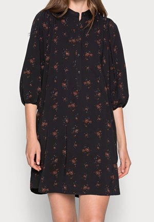 Kobieta ubrana w czarną sukienkę do kolan z brązowym kwiatowym wzorem, z rękawami o długości za łokieć zakończonymi bufkami oraz zapinanym na guziki kołnierzykiem.