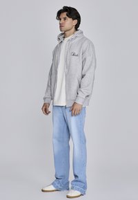 Sivá mikina s predným vreckom, kapucňou so šnúrkou a vyšitým logom. Nosená cez bielu košeľu s svetlo modrými džínsami a teniskami.