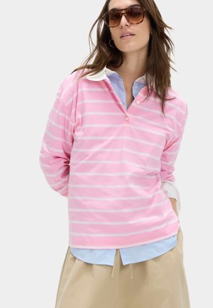 Frau trägt ein übergroßes Rugby-Shirt mit rosa-weißen Streifen über einem blauen Hemd mit Kragen, einem beigen Rock und Schildpatt-Sonnenbrillen.