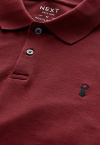 Bordo polo majica od teksturiranog materijala, s tri siva dugmeta i crnim izvezenim logom jastoga na grudima.