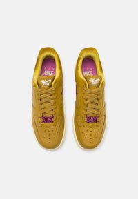 Paire de baskets Nike Air Force 1 jaune moutarde avec des charms violets « Just Do It » sur les lacets et des semelles intérieures roses, vue de dessus.