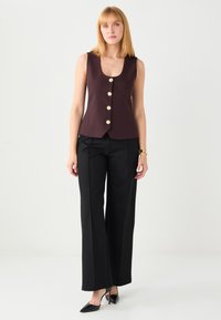 Donkerrood mouwloos vest met vijf gouden knopen, gecombineerd met zwarte broek met wijde pijpen en punten zwarte hakken. Glad stoftextuur.